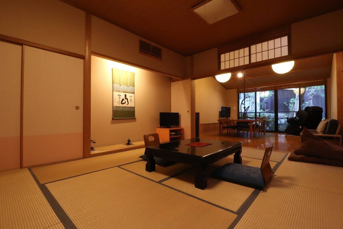 A traditional Japanese room with tatami flooring, a low table, a television, and a view of a garden through large windows.