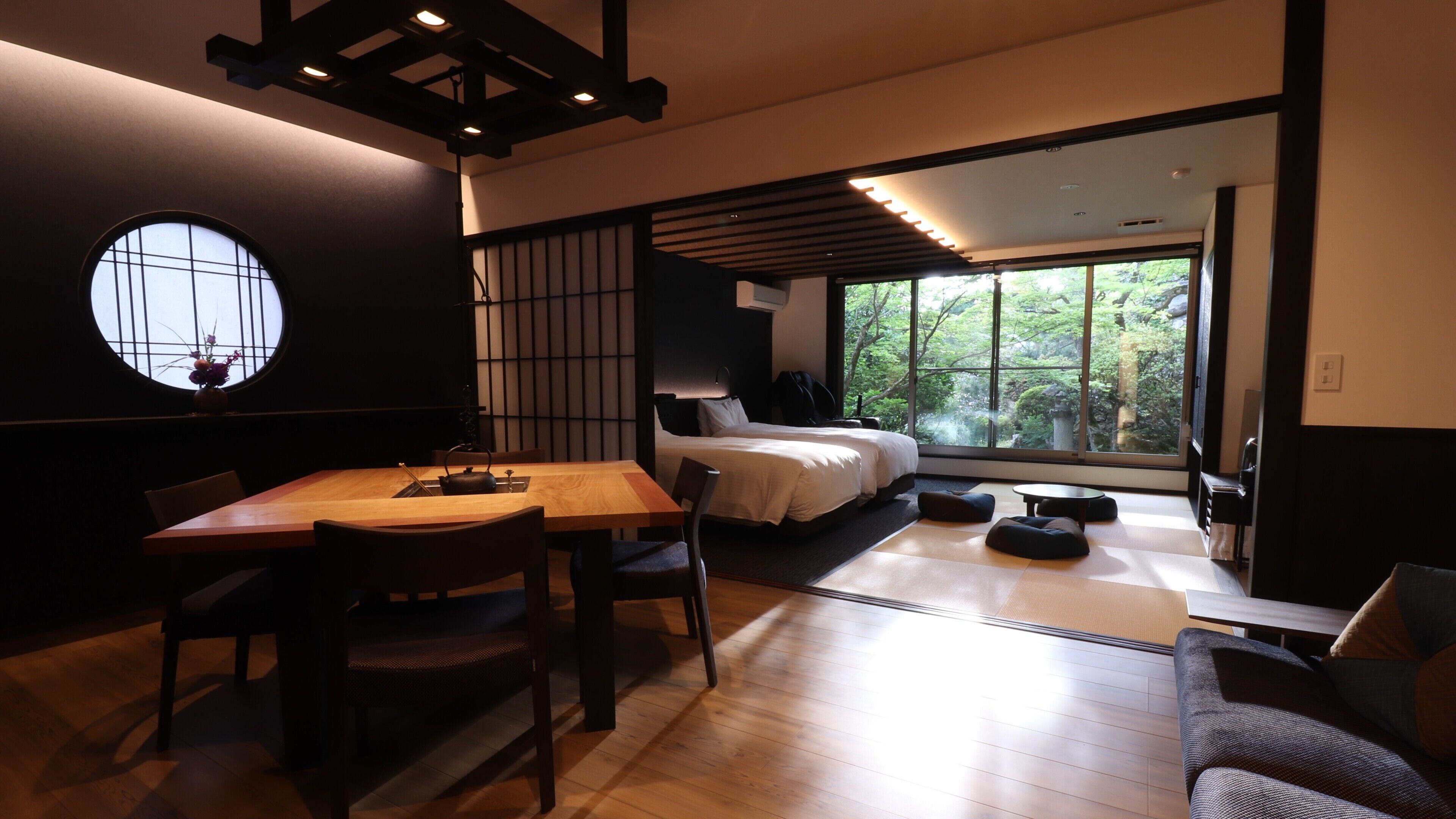 A modern hotel room with a wooden dining table, a sofa, a bed, and a large window offering a view of greenery.