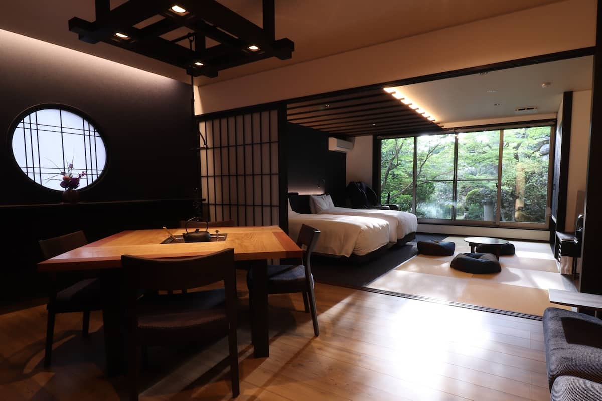 Una habitación de hotel moderna con una mesa de comedor de madera, un sofá, una cama y un ventanal con vistas a la vegetación.