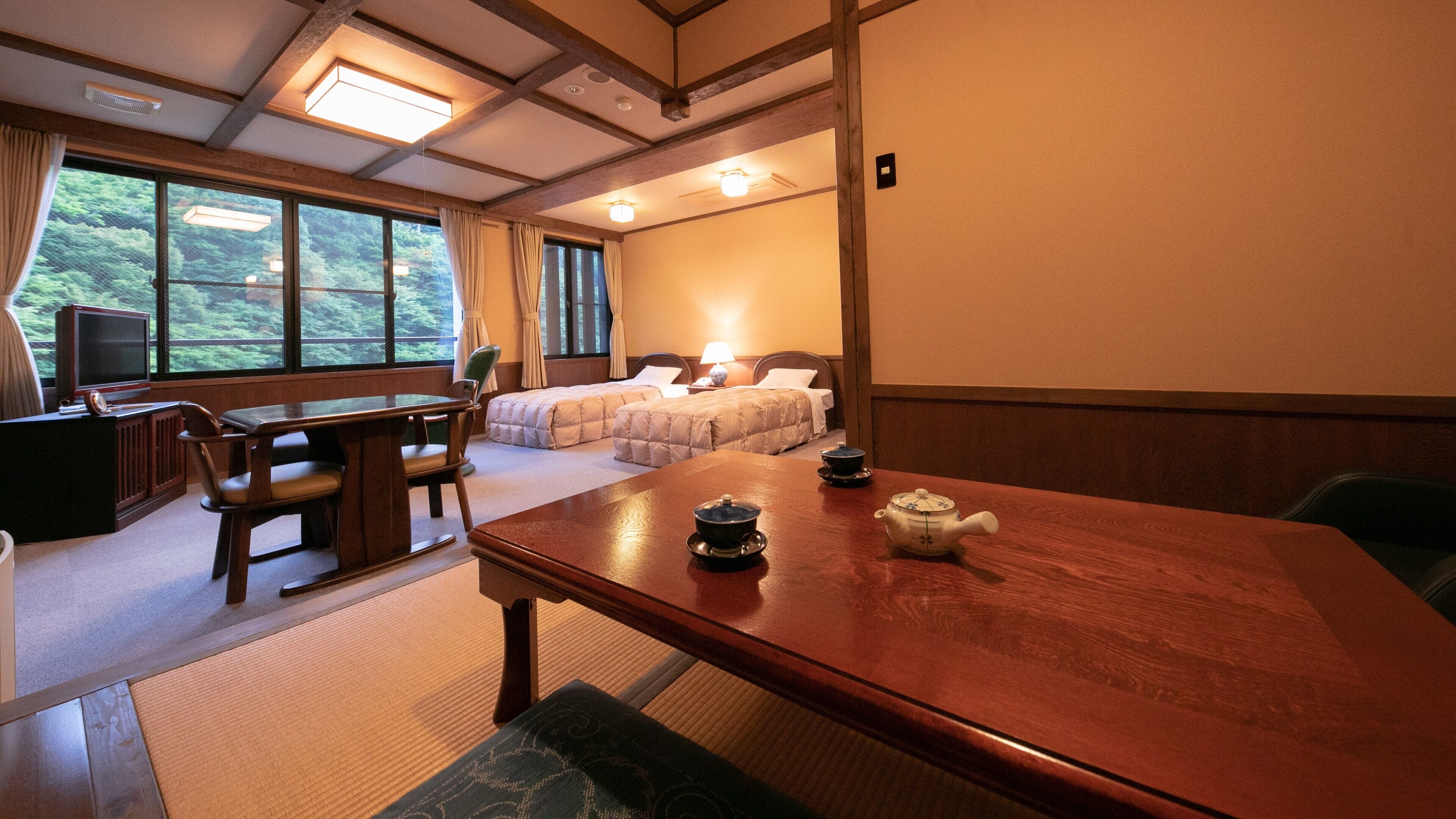 A hotel room with a wooden dining table, two beds, a television, and a view of greenery.