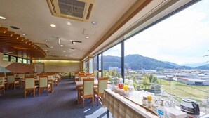 A dining area with tables and chairs, a large window with a view of buildings and mountains, and a counter with various items. - Hotel Areaone Wadayama (Asago)