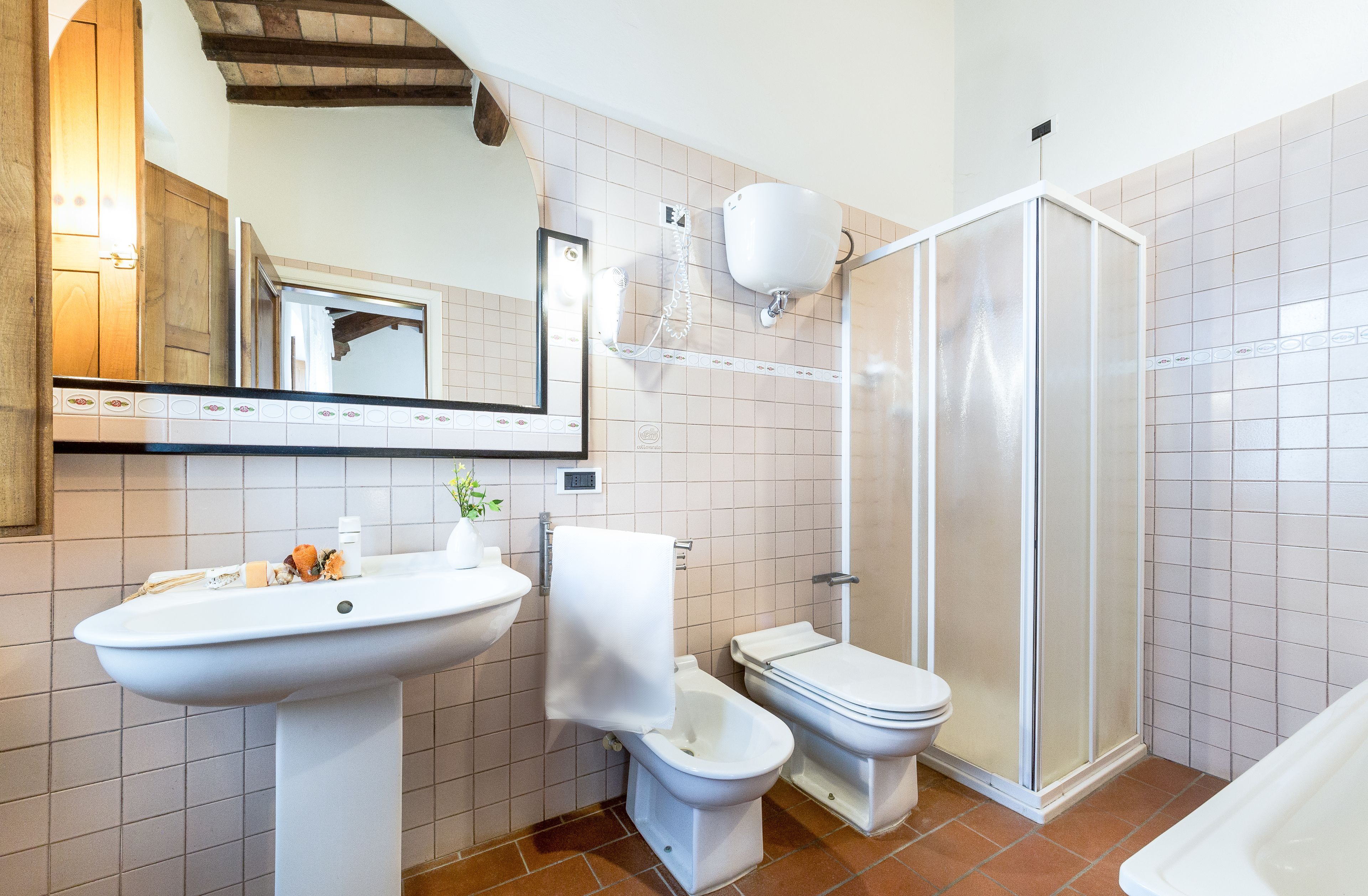 Un baño con lavamanos pedestal, dos inodoros, cabina de ducha y espejo sobre el lavamanos.
