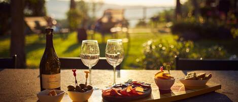 A table with a bottle of wine, two glasses, a bowl of olives, a plate of food, and a bowl of bread, set against a scenic outdoor view.