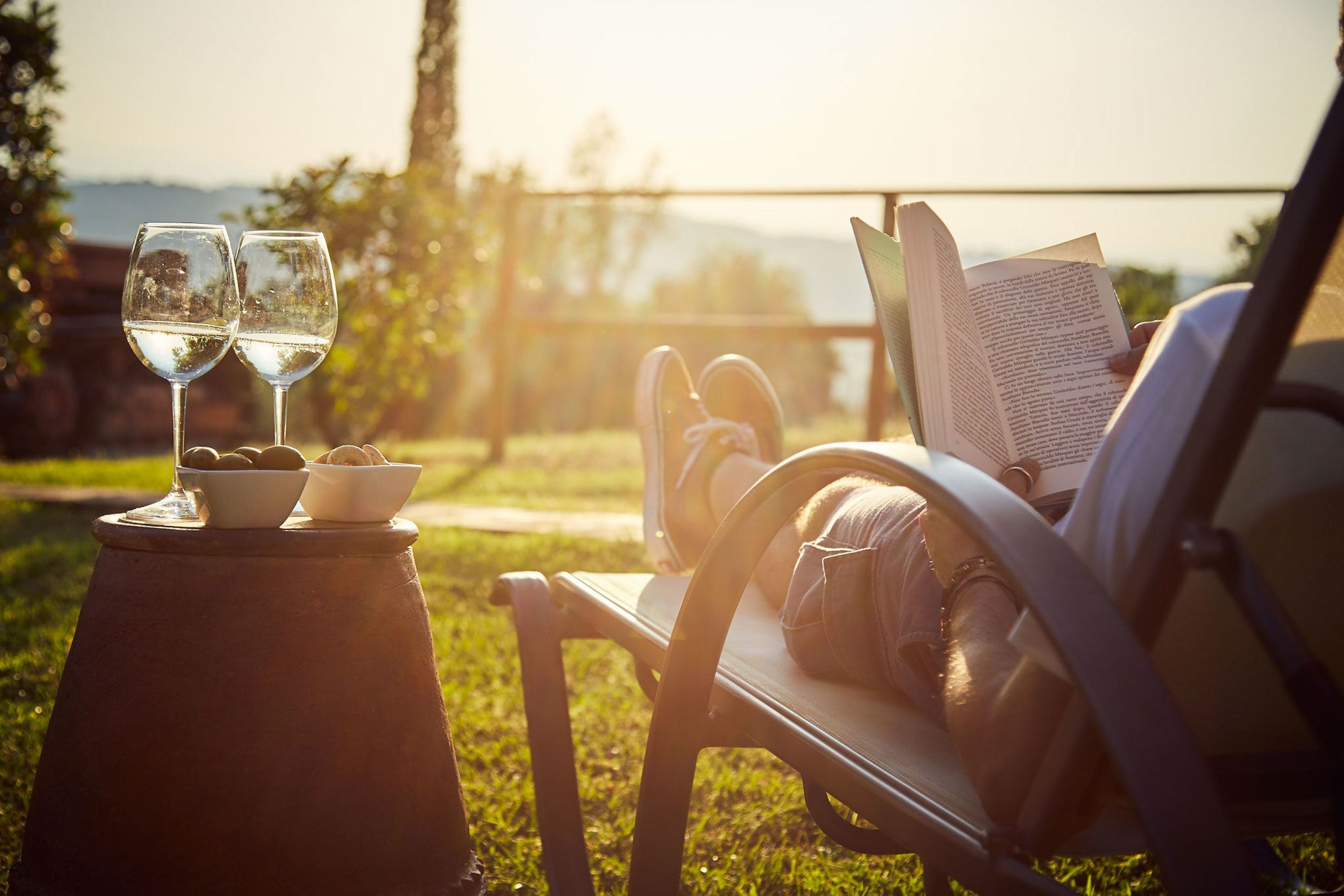 A person relaxing on a chair with a book and two glasses of wine on a stool.
