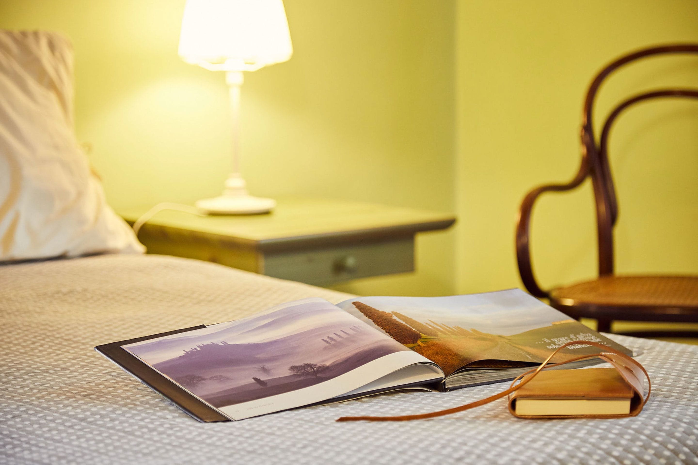 A bed with a book and a magazine, a nightstand with a lamp, and a chair in a room with yellow walls.