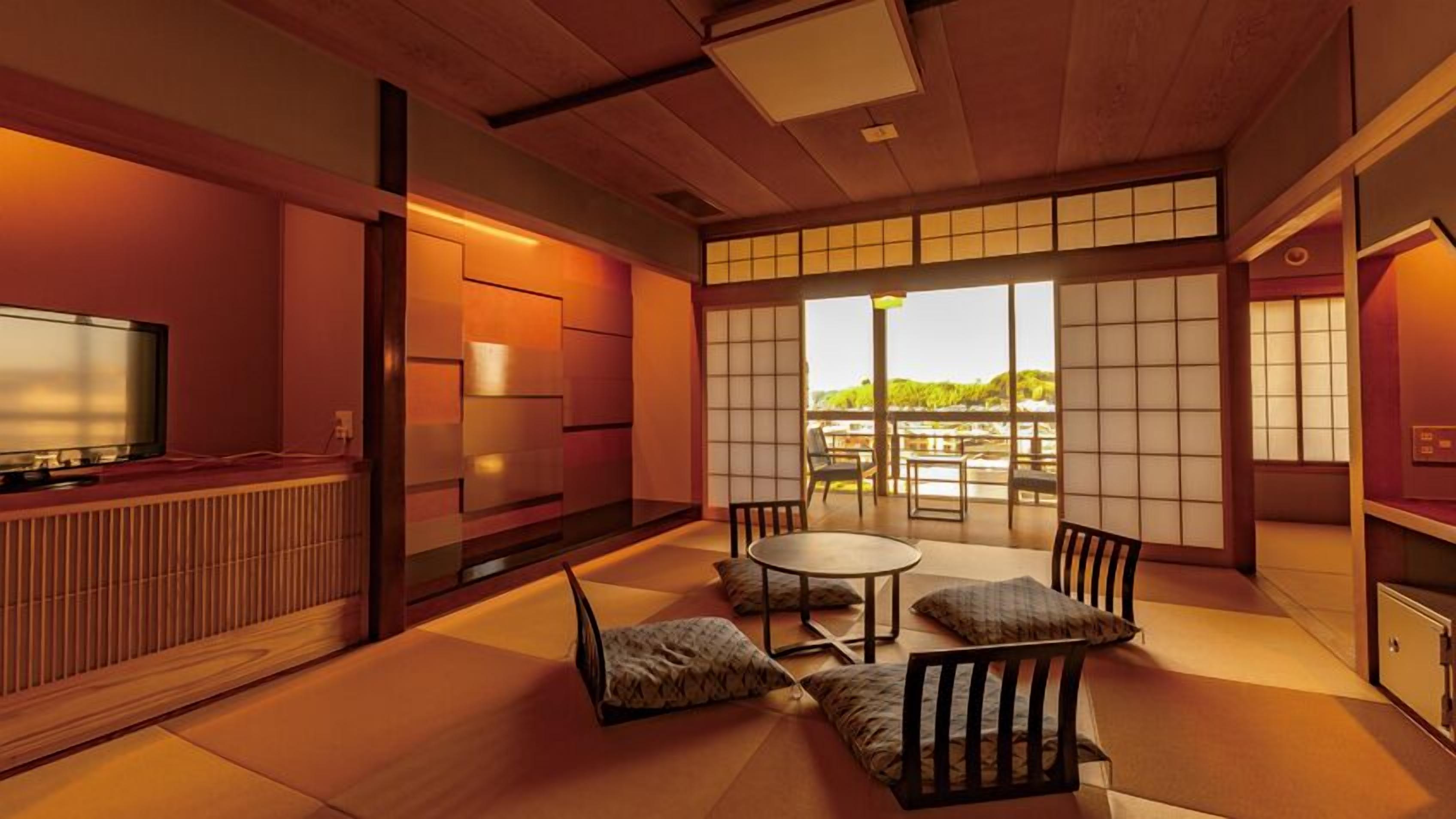 Una habitación tradicional japonesa con suelo de tatami, una mesa baja, sillas, televisor y ventanas amplias con vistas a la vegetación.