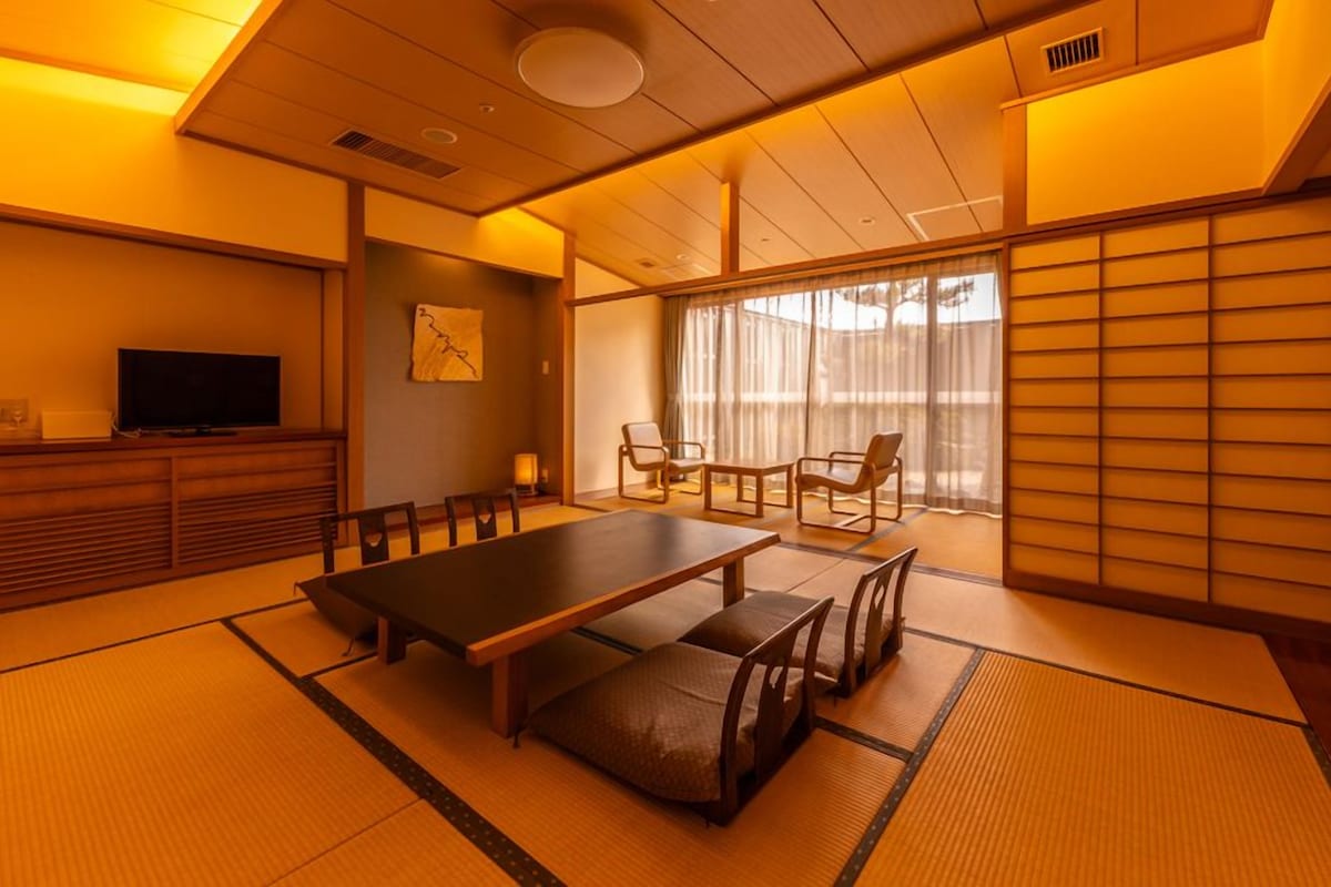 A traditional Japanese room with tatami flooring, a low table, chairs, a television, and sliding doors.