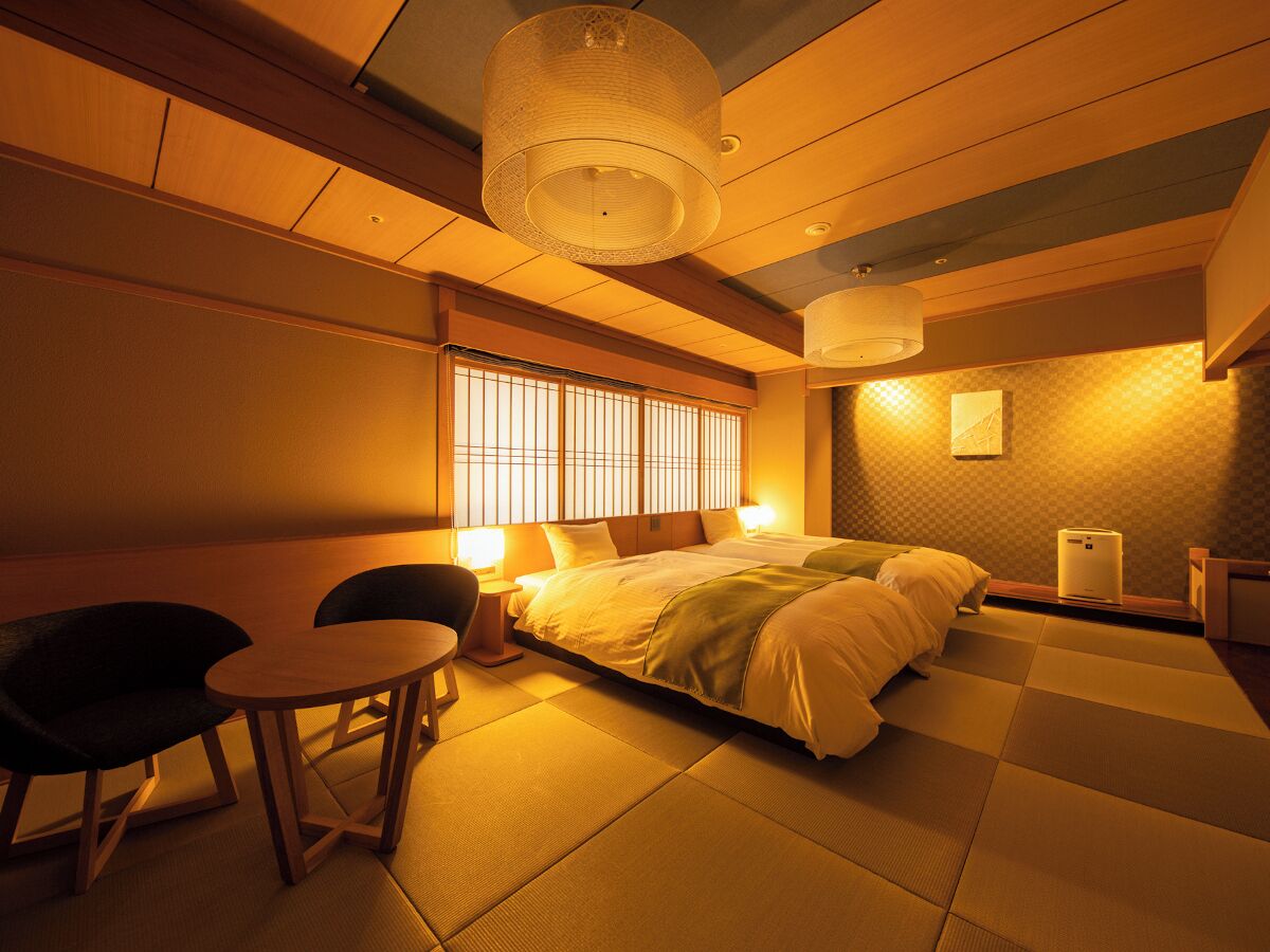 Habitación de hotel con cama, una mesita, dos sillas, ventana con persianas y una lámpara en el techo.