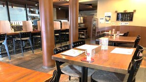 L’intérieur d’un restaurant présente des tables et des chaises en bois, un bar avec des bouteilles exposées et un tableau affichant le menu sur le mur.