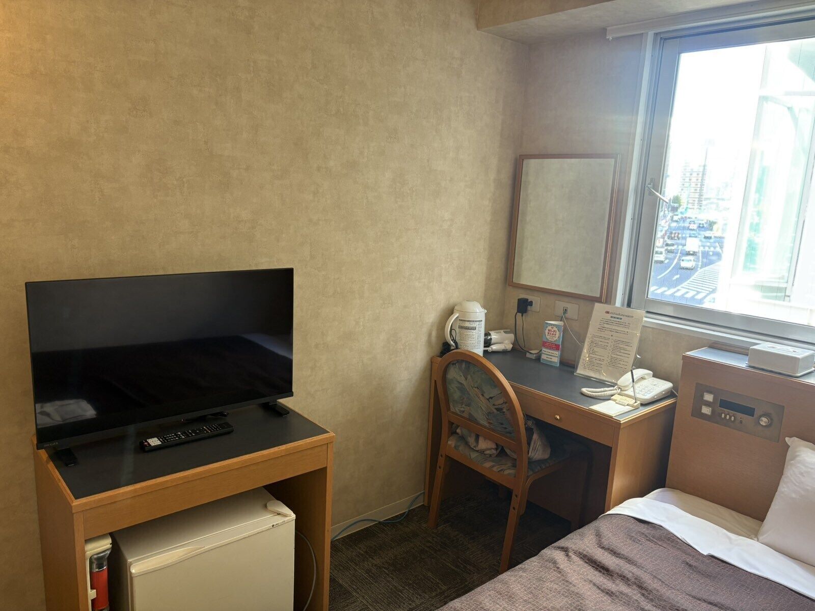 Une chambre d’hôtel équipée d’un lit, d’un bureau avec un ordinateur, d’une chaise, d’une télévision et d’une fenêtre donnant sur la ville.