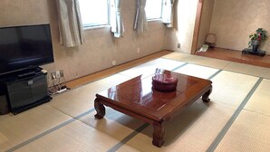 Une pièce avec un sol recouvert de tatamis, une table basse en bois sur laquelle se trouve un récipient rouge, un téléviseur posé sur un meuble et une fenêtre avec des rideaux.