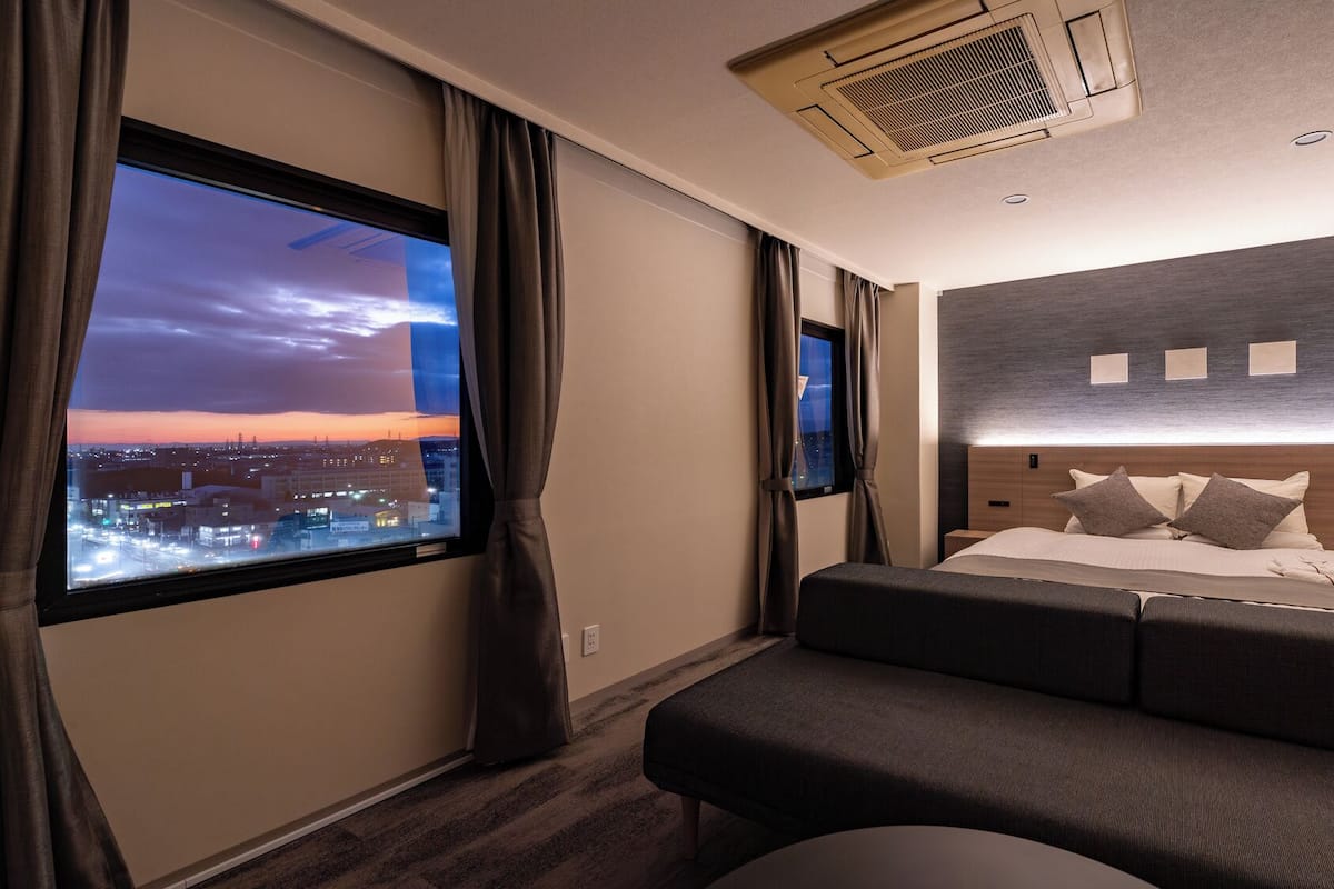 A hotel room with a large window, a bed with a headboard, a bench, and a wall-mounted TV.