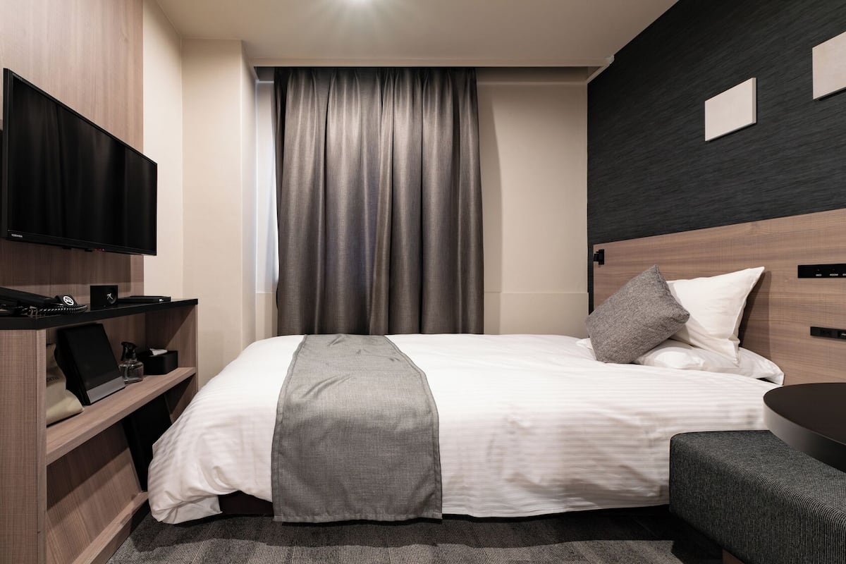 A modern hotel room with a bed, a desk with a flat-screen TV, a chair, and a control panel on the wall.