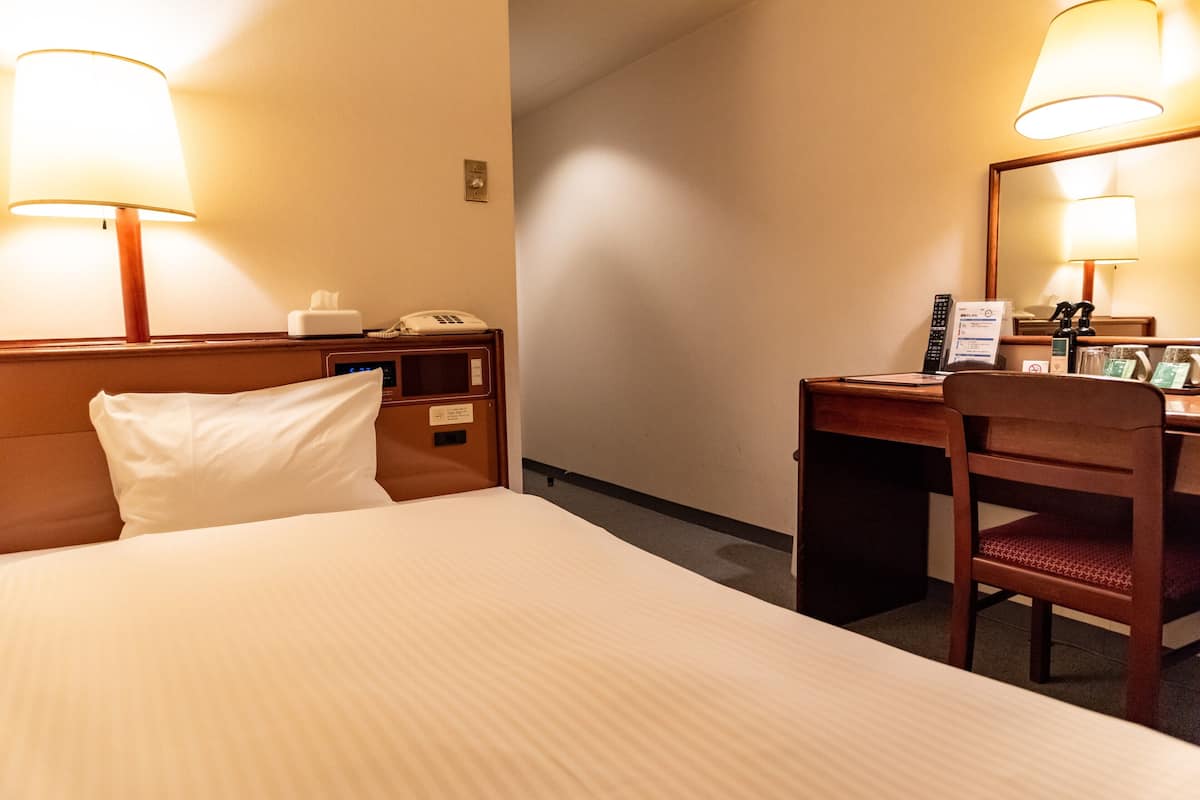 A hotel room with a bed, bedside table, lamp, desk, chair, and a mirror.