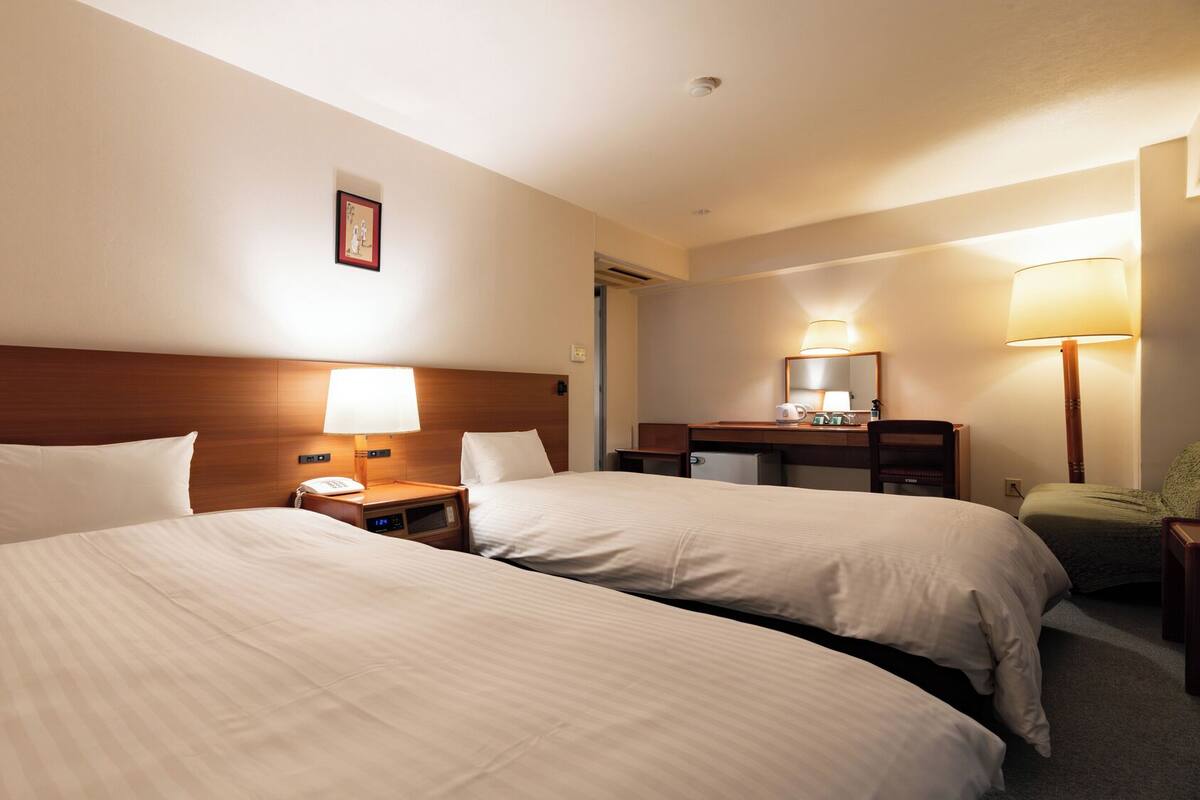 A hotel room with two beds, a desk, a chair, a lamp, and a small table with a lamp.