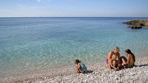 Une plage de galets avec une eau claire et des bateaux qui se dessinent à l’horizon.