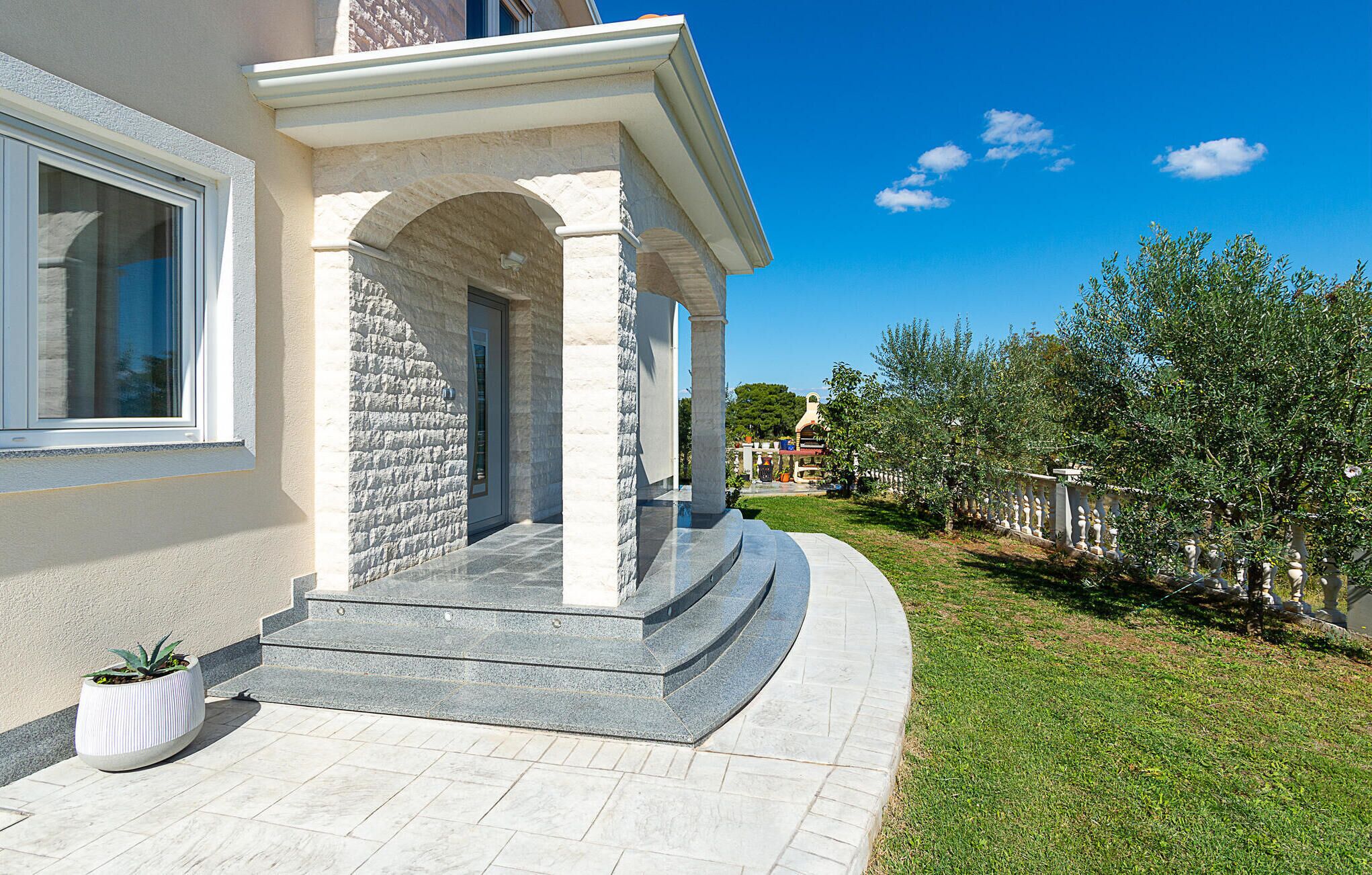 A modern building with a stone facade, a covered entrance, and a well-maintained garden with olive trees.