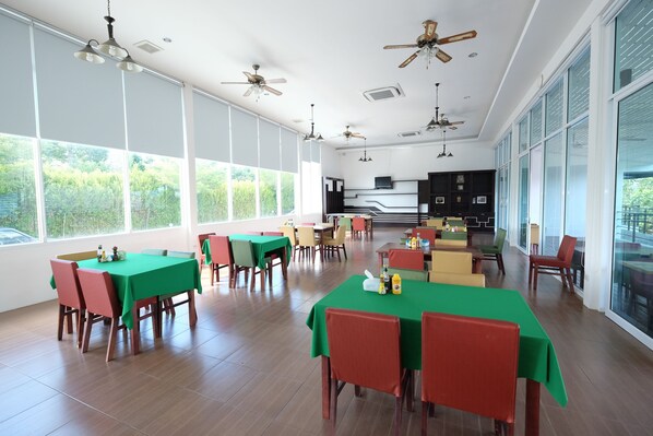 Un comedor amplio con varias mesas y sillas, ventanas grandes y ventiladores de techo.