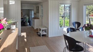 Tilava huone, jossa on puulattia, ruokapöytä tuoleineen, pieni pöytä kenkiä varten ja suuri ikkuna, josta avautuu näkymä vehreään maisemaan.