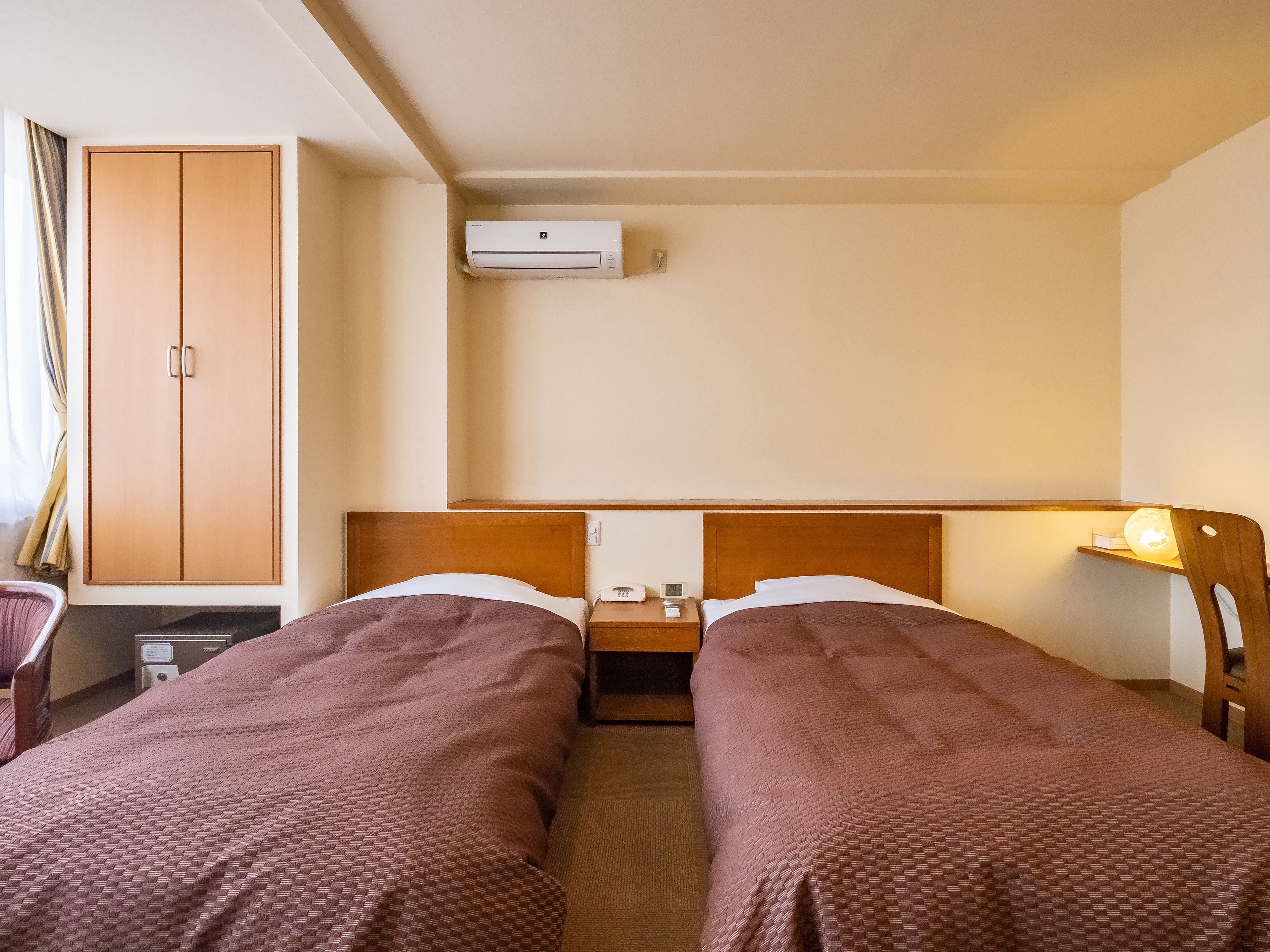 Habitación de hotel con dos camas, un armario, un equipo de aire acondicionado, una mesita de noche con lámpara y un escritorio con silla.