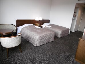 Habitación de hotel con dos camas, una silla, una mesita pequeña y una mesita de noche con lámparas.