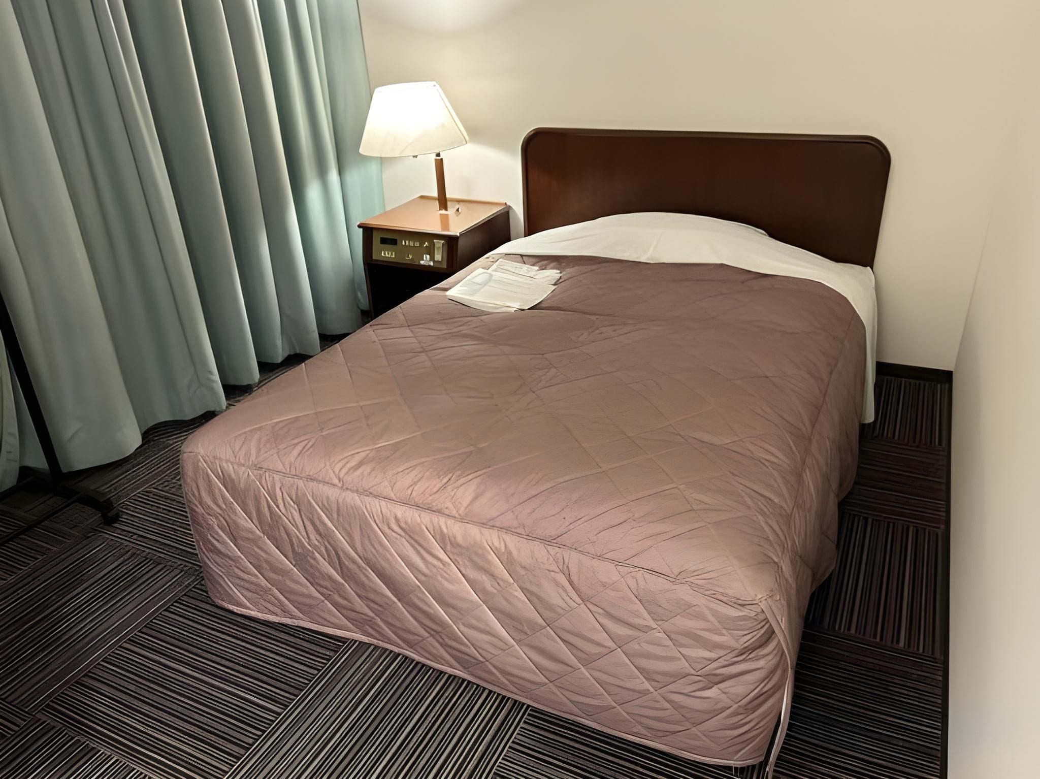 A hotel room with a bed, a nightstand, a lamp, and a window with curtains.