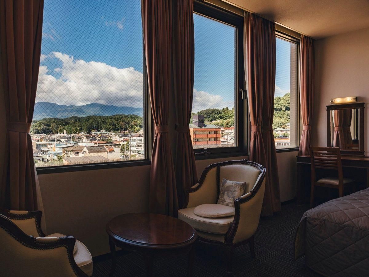 Habitación de hotel con cama, una silla, una mesita y vistas a un paisaje urbano a través de amplios ventanales.