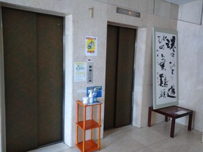 Elevador com placa indicando o primeiro andar, uma pequena mesa com cadeira e uma prateleira com produtos de limpeza.