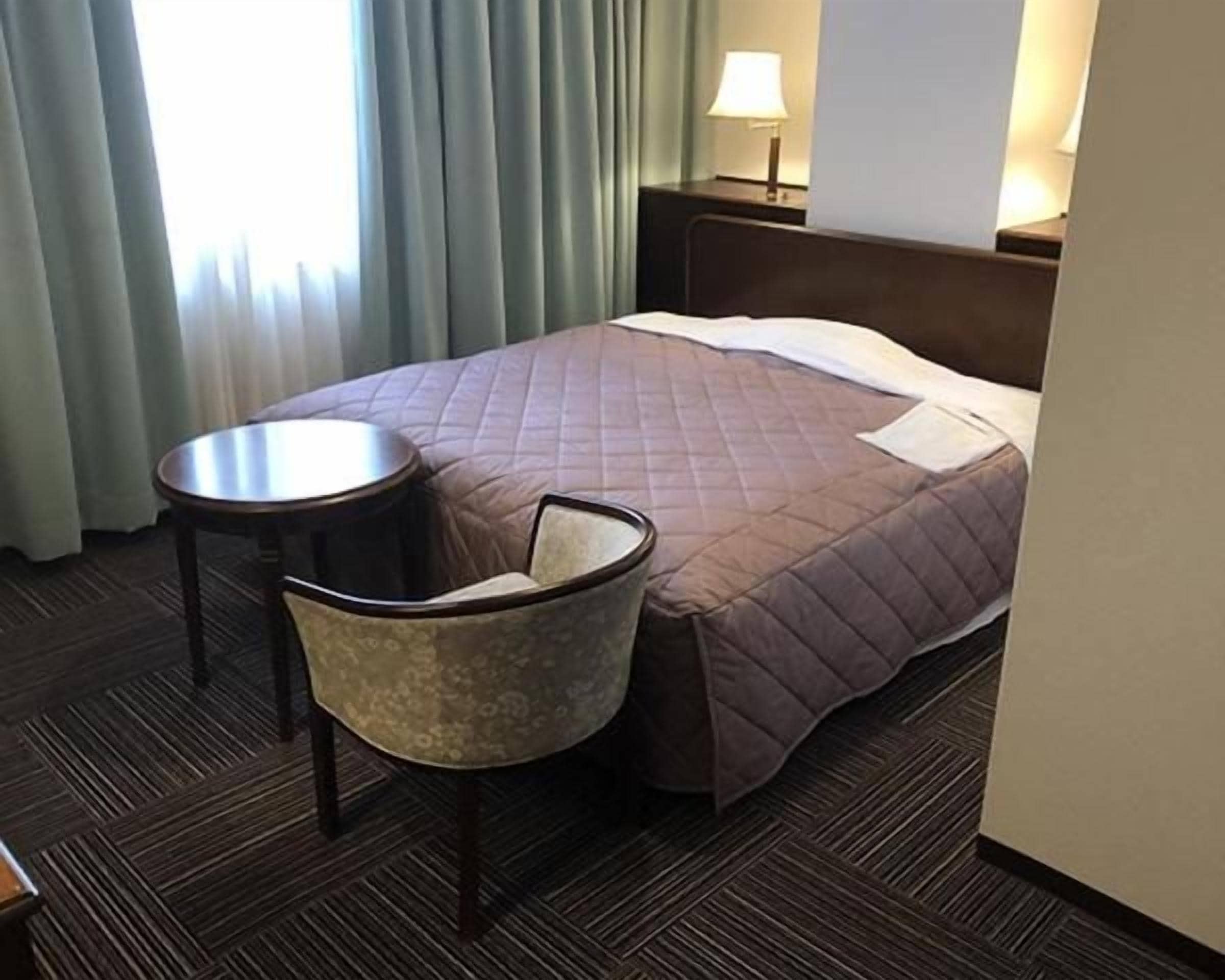 A hotel room with a bed, a chair, a small table, and a lamp.