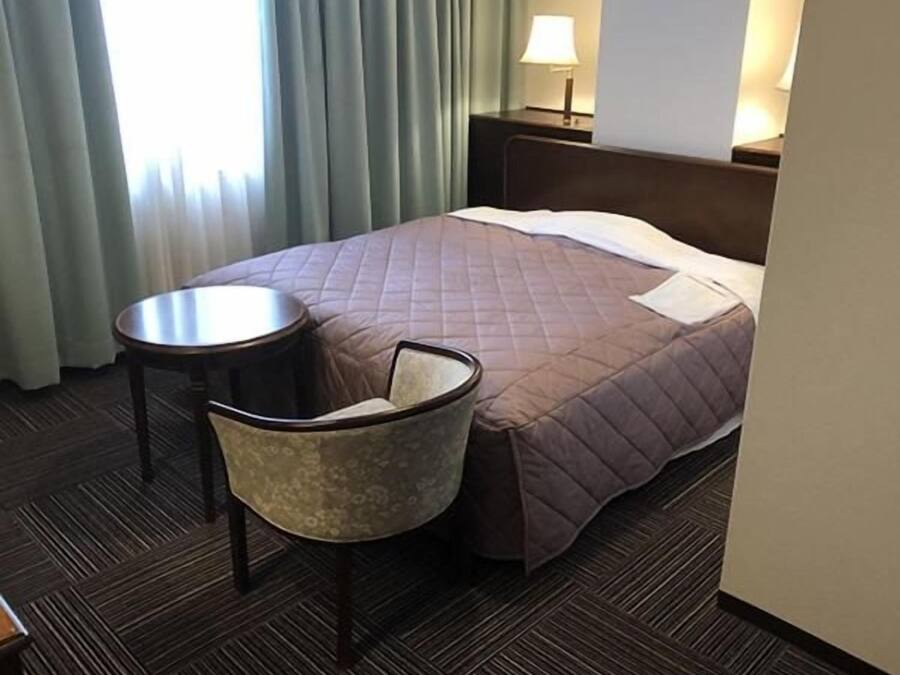 A hotel room with a bed, a chair, a small table, and a lamp.