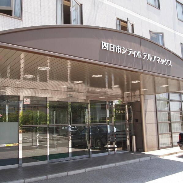 Entrada de hotel con fachada de vidrio, área de recepción y un letrero que indica un DEA.