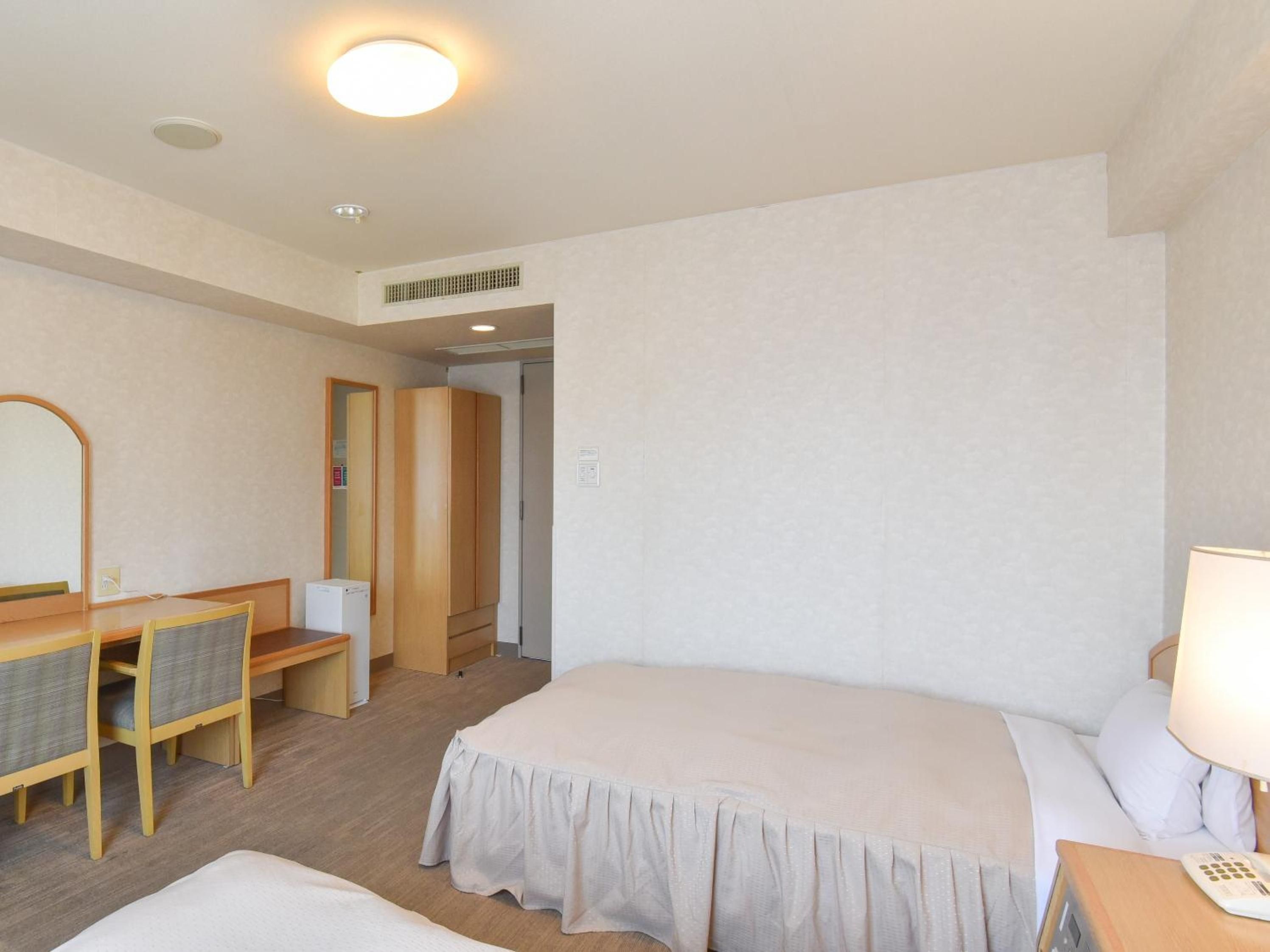 A hotel room with a bed, a desk with a chair, a mirror, and a bedside table with a lamp.