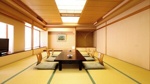Una tradizionale stanza con tatami, un tavolo lungo, tatami, sedie di legno, un quadro appeso al muro e un televisore a sinistra.