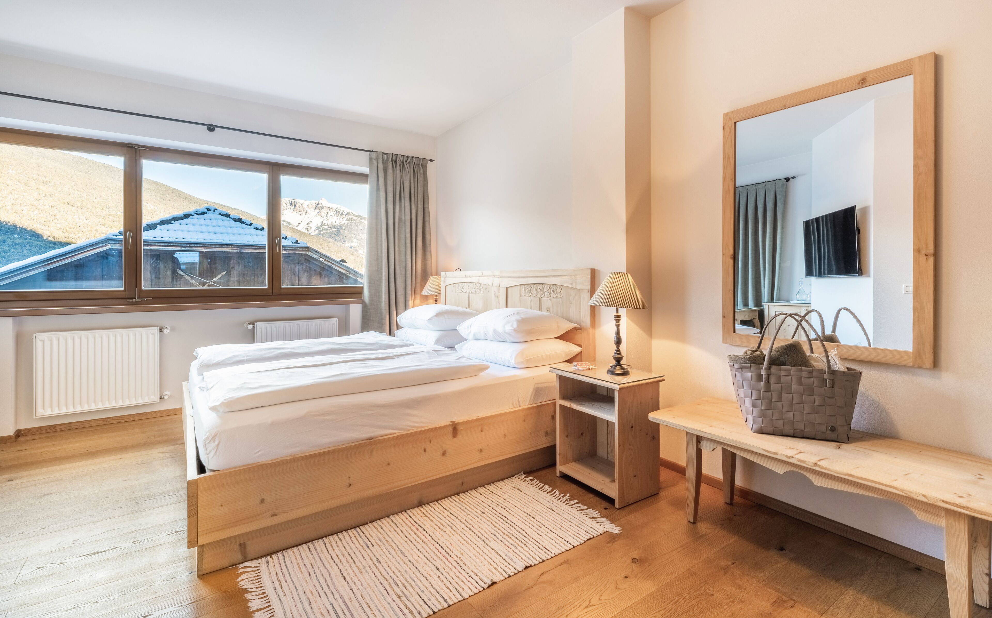 A bedroom with a wooden bed, a bedside table with a lamp, a mirror, and a view of mountains through large windows.