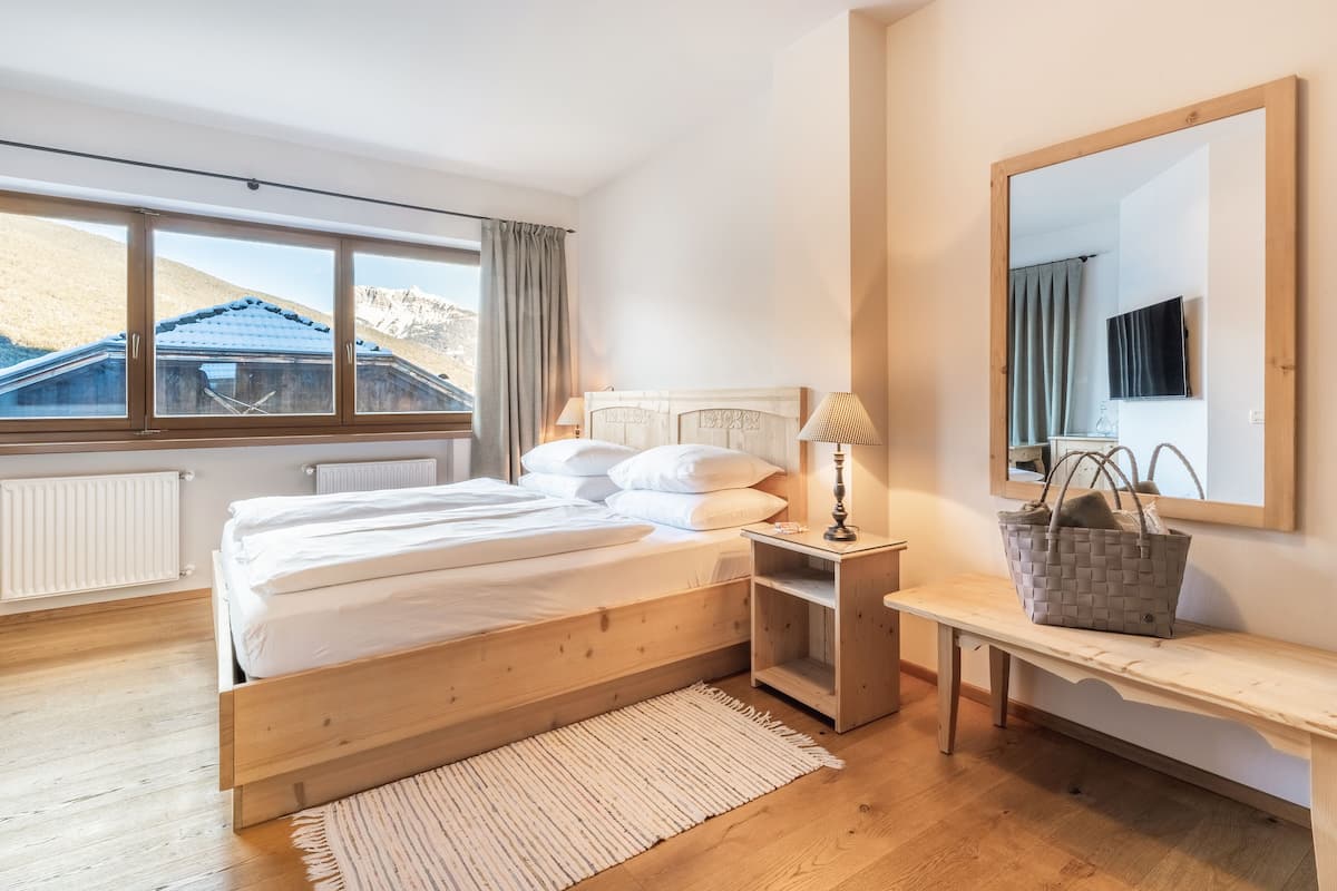 Un dormitorio con una cama de madera, una mesita de noche con lámpara, un espejo y vistas a las montañas a través de amplios ventanales.