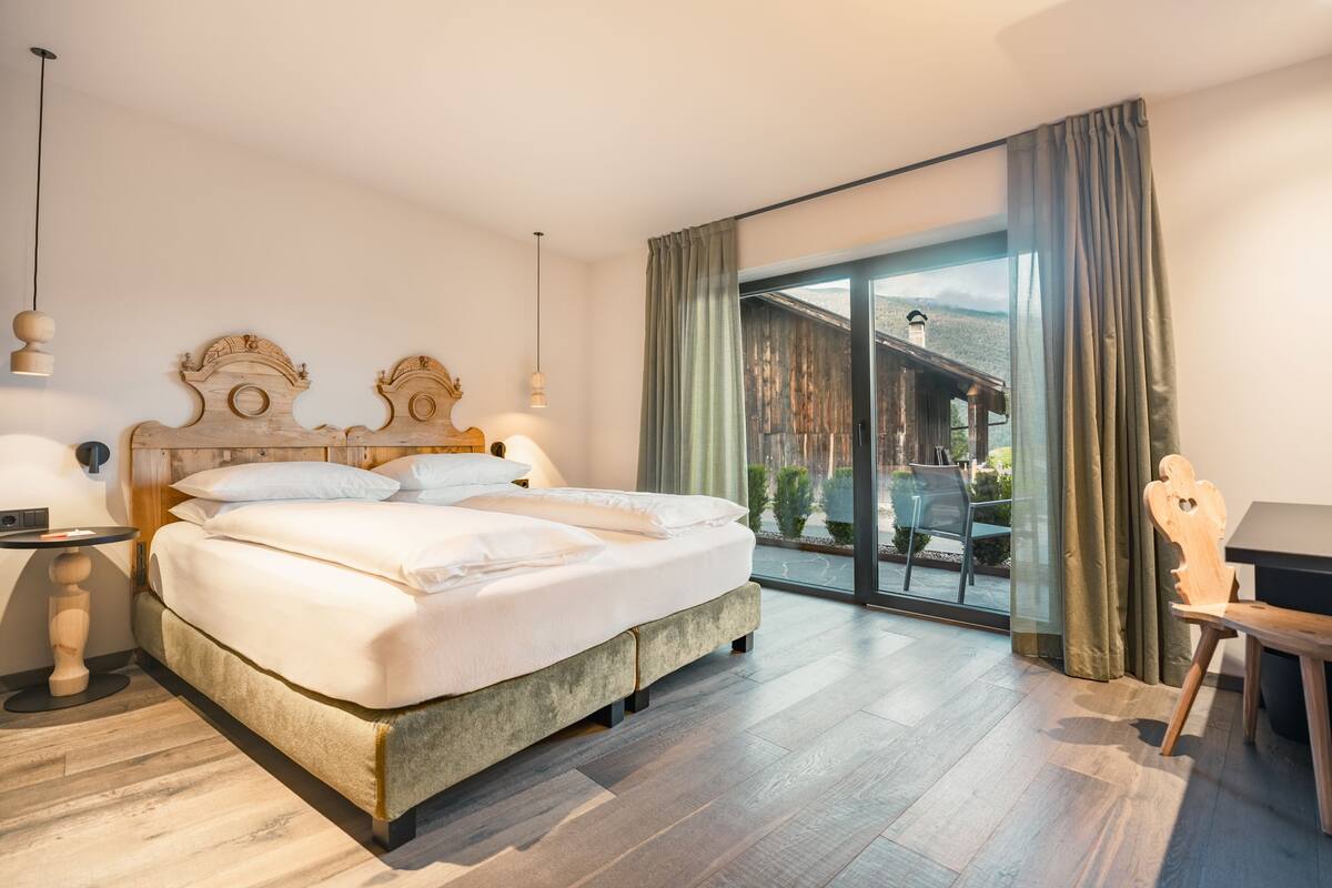 Una habitación de hotel moderna con una cama grande, un cabecero de madera, una mesita, una silla y vistas a una cabaña de madera a través de una puerta corrediza de vidrio.