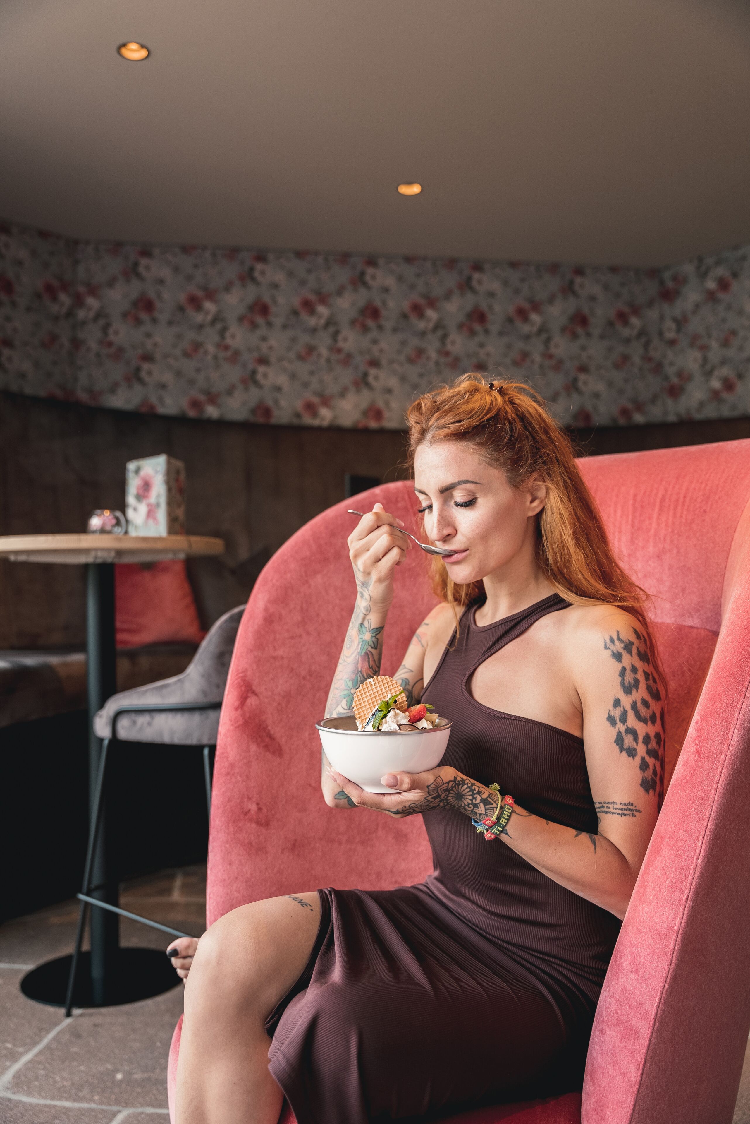 A person sitting on a red chair in a room with floral wallpaper, holding a bowl of food.