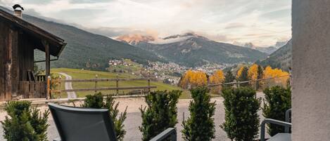 A terrace with outdoor seating, overlooking a mountainous landscape with autumn foliage and a distant village.