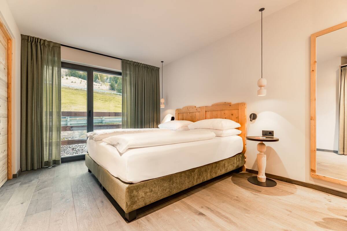 Un dormitorio moderno con una cama grande, cabecera de madera, mesita de noche con lámpara y vista a un espacio verde a través de una puerta corrediza de vidrio.