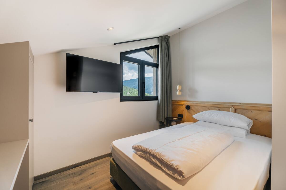 Un dormitorio con una cama, un televisor montado en la pared, una ventana con cortinas y una mesita de noche con lámpara.