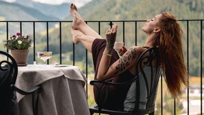 A person relaxing on a balcony with a table, potted plants, and a view of mountains.