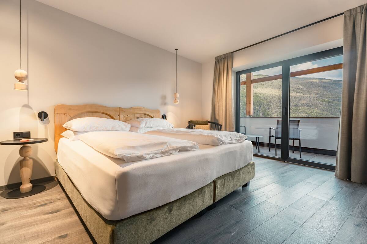 Habitación de hotel moderna con una cama grande, una mesita pequeña y un balcón con vistas a las colinas.