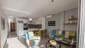 A modern living room with a sofa, armchair, and dining table. There is a kitchen in the background with a refrigerator, oven, and sink.