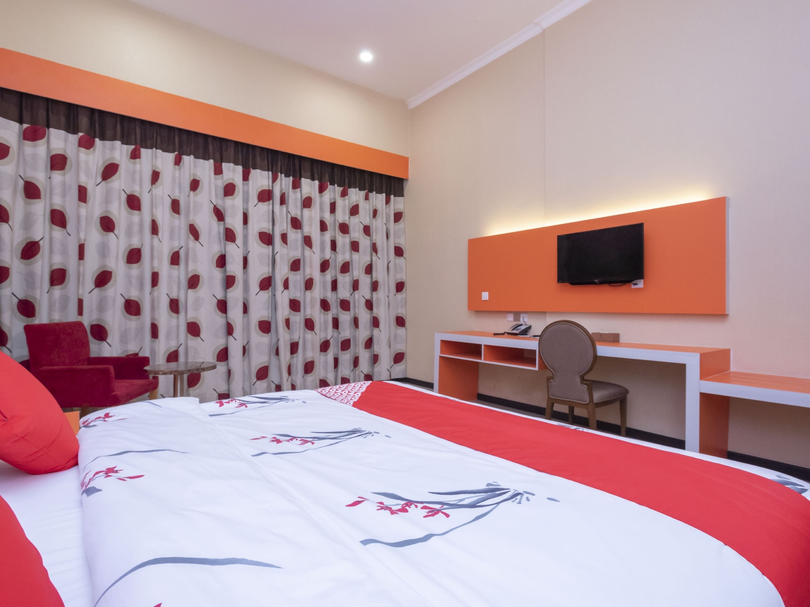Una habitación de hotel con cama, escritorio, silla, televisor y un sillón rojo.