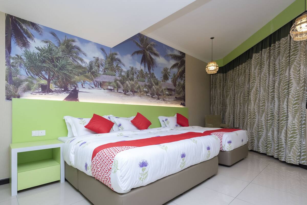 A double bed with a floral bedspread and red pillows, a green nightstand, a large tropical mural on the wall, and a hanging light fixture.