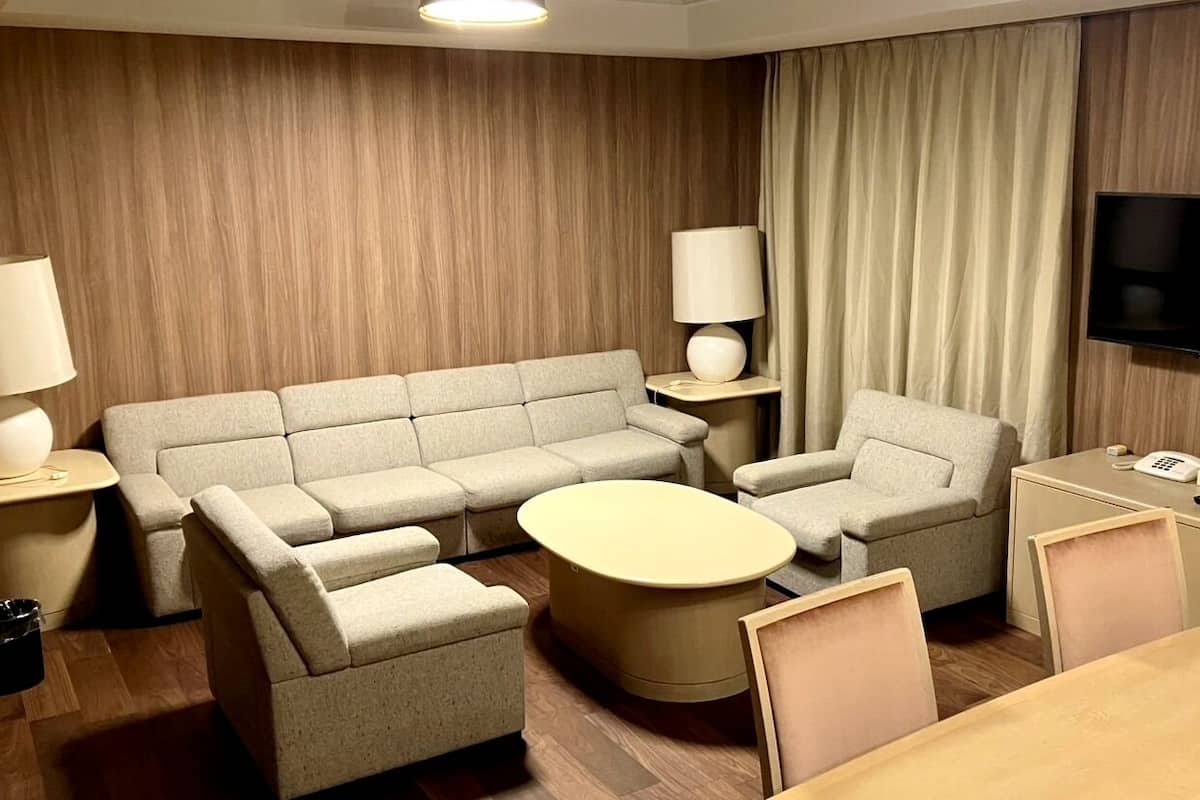 A modern hotel room with a sofa set, a round coffee table, a TV, and a desk with a phone.