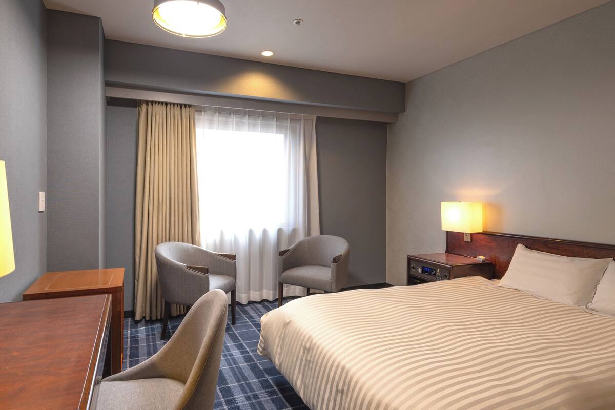 A hotel room with a bed, two armchairs, a desk, a lamp, and a nightstand with a lamp.