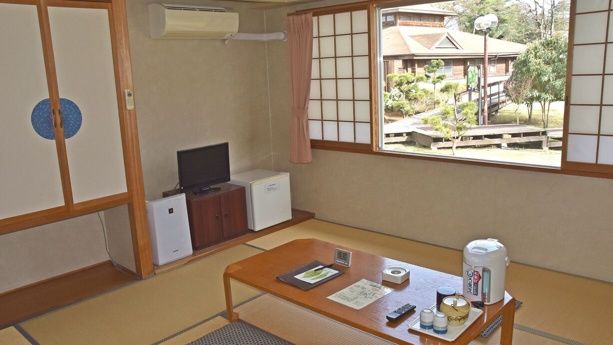 Une pièce de style japonais traditionnel, avec un sol recouvert de tatamis, une télévision, un petit réfrigérateur, une table en bois avec une télécommande, une bouilloire et une vue sur un quartier résidentiel.