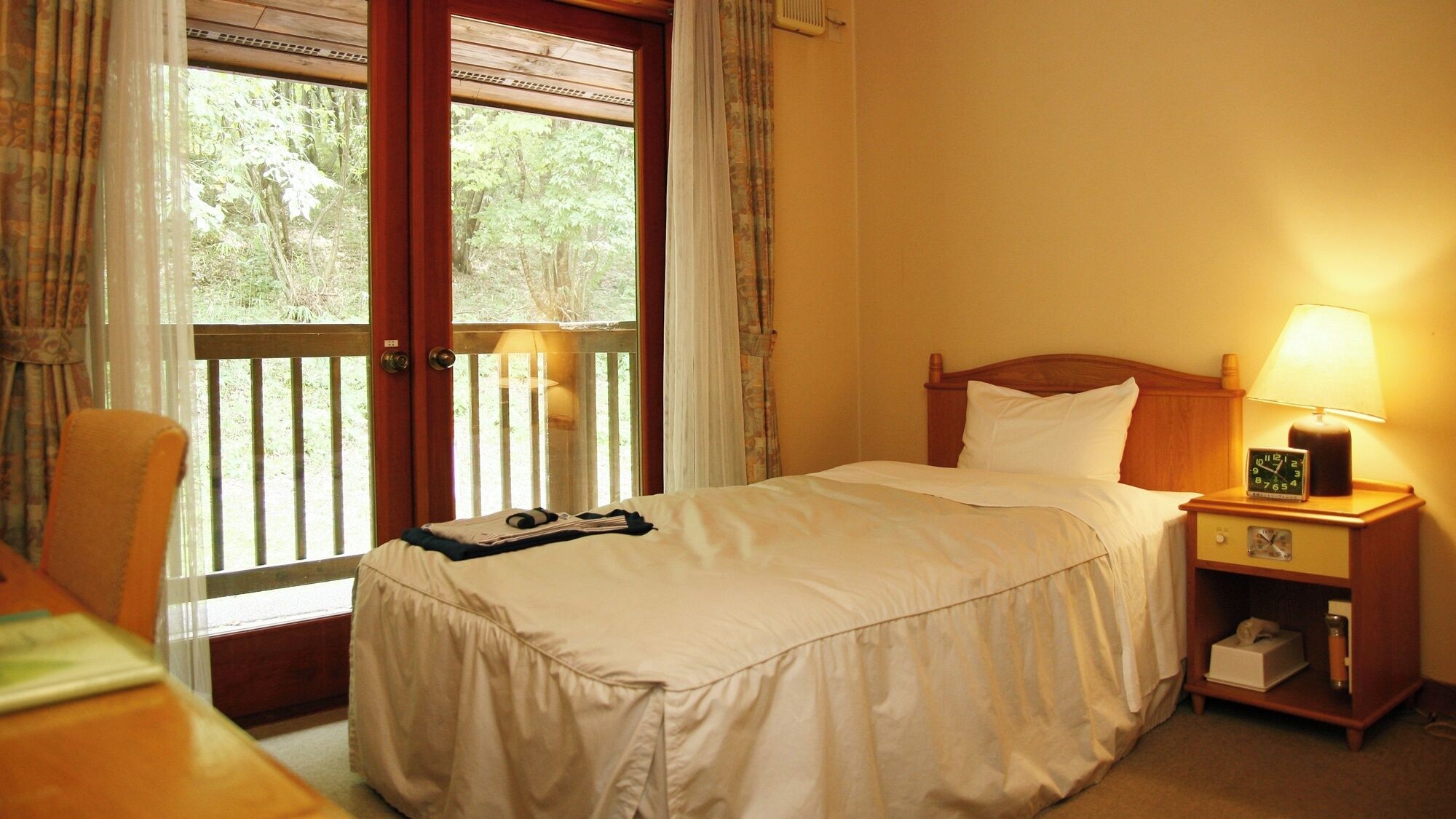 A hotel room with a bed, a bedside table with a lamp, a nightstand, a chair, and a window with curtains.