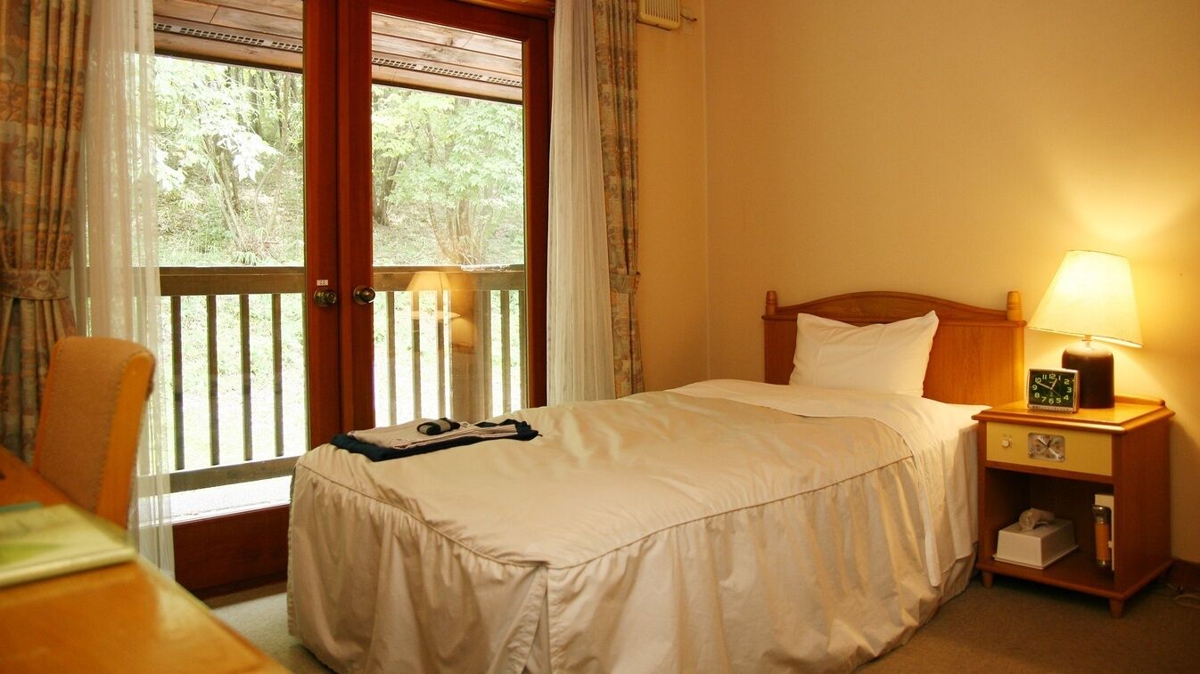 Une chambre d’hôtel avec un lit, une table de chevet avec une lampe, une chaise et une fenêtre avec des rideaux.